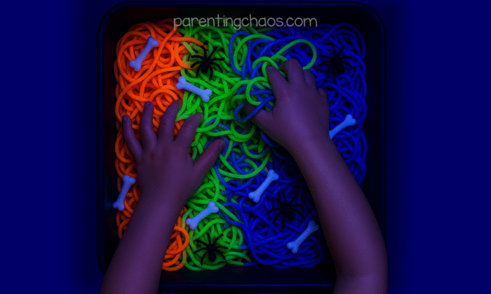 A child’s hands playing in the glowing spaghetti sensory bin, squishing the colorful noodles and plastic bones under black light.