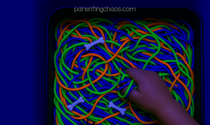 A close-up of a child’s hand digging into a tangle of glowing Halloween spaghetti noodles with bones scattered throughout.