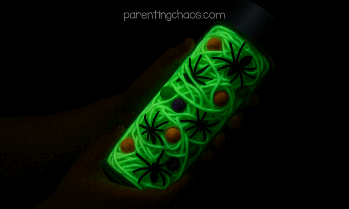 The same sensory bottle glowing bright green in the dark, with spiders and colorful balls visible inside, held in a child’s hands.