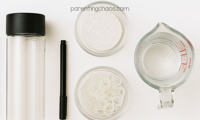 Overhead view of glowing ghost sensory bottle supplies — clear bottle with black lid, black marker, glitter, water, and clear rubber bands arranged neatly on a white background.