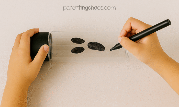 Child drawing a ghost face on a clear sensory bottle using a black permanent marker.