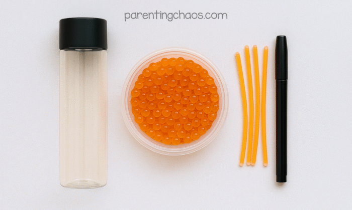 Supplies for a glowing pumpkin sensory bottle, including an empty clear bottle with a black lid, orange water beads in a bowl, orange glow sticks, and a black marker.