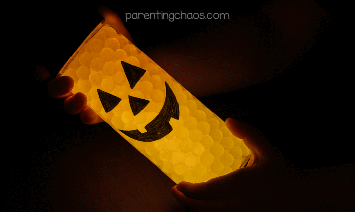 Child holding a glowing pumpkin sensory bottle in the dark, showing the lit jack-o’-lantern face.