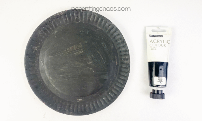 A paper plate painted black next to a tube of black acrylic paint.