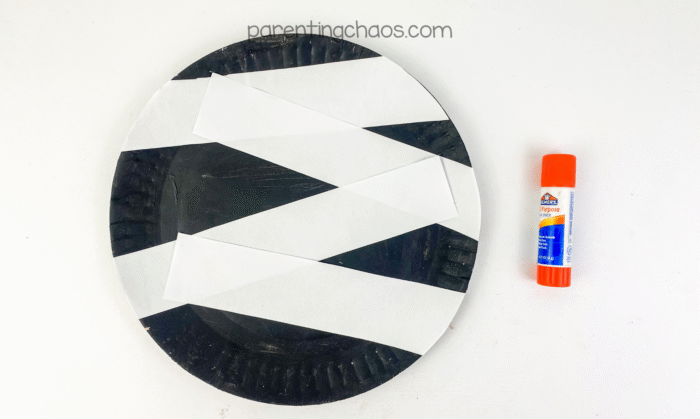 A black painted paper plate with white strips glued diagonally across it and a glue stick beside it.