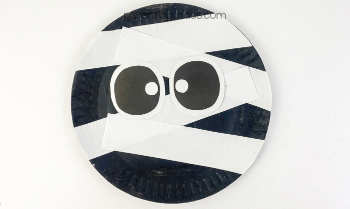 A finished mummy paper plate craft with white paper wraps and large cartoon eyes on a black background.