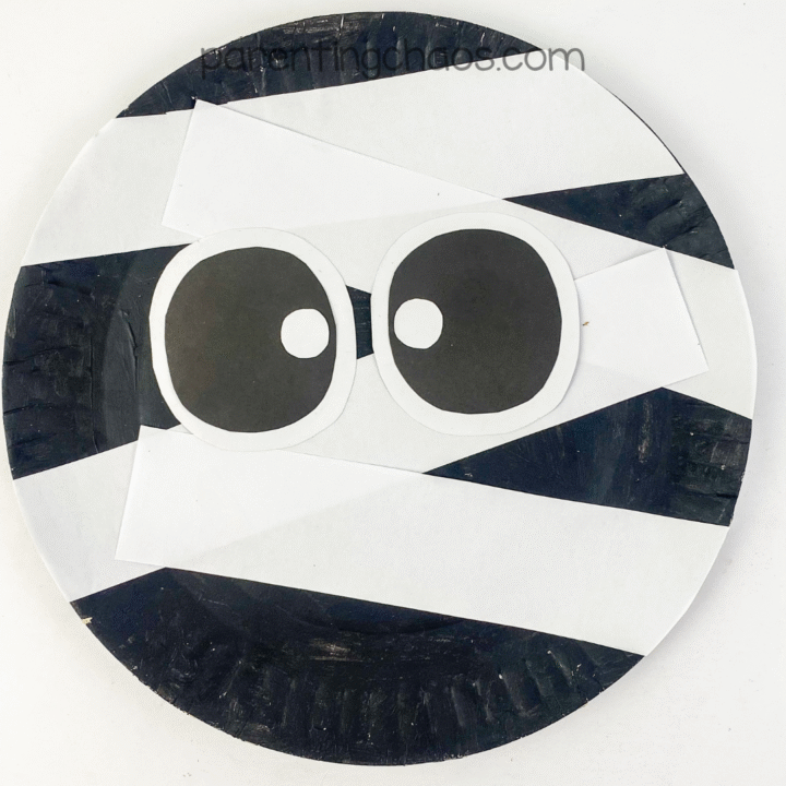 A finished mummy paper plate craft with white paper wraps and large cartoon eyes on a black background.