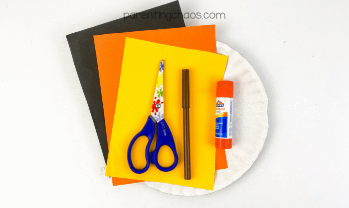 Crafting supplies for owl paper plate craft: black, orange, and yellow construction paper, preschool scissors with floral handles, brown marker, Elmer’s glue stick, and a white paper plate.