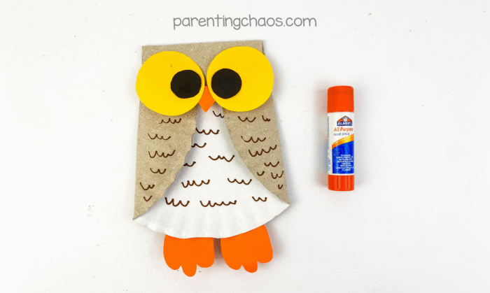Large yellow and black owl eyes glued to the folded paper plate body with an orange beak, glue stick beside it.
