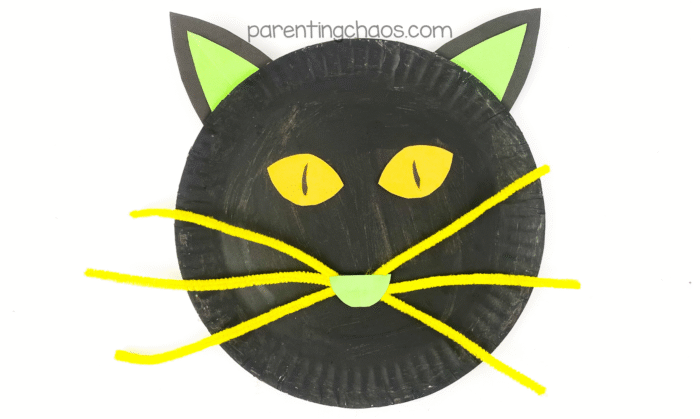 Finished black cat paper plate craft with yellow eyes, green nose, green and black ears, and long yellow pipe cleaner whiskers — completed Halloween craft.