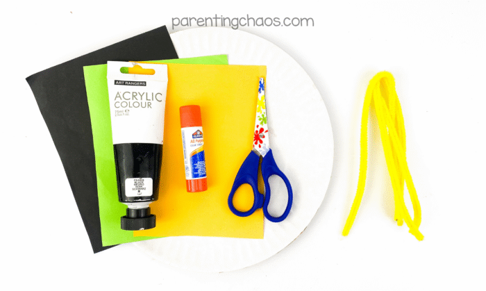 Overhead view of black, green, and yellow construction paper, black acrylic paint, glue stick, colorful scissors, yellow pipe cleaners, and a white paper plate — all the supplies needed for a black cat paper plate craft.