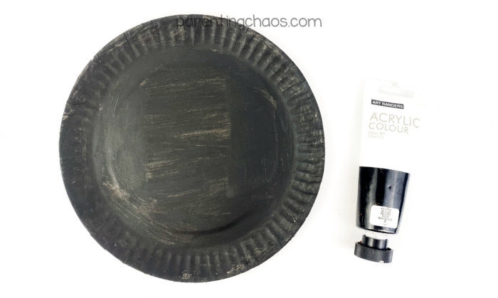 Paper plate painted solid black with a tube of black acrylic paint next to it, showing the base for the black cat craft.