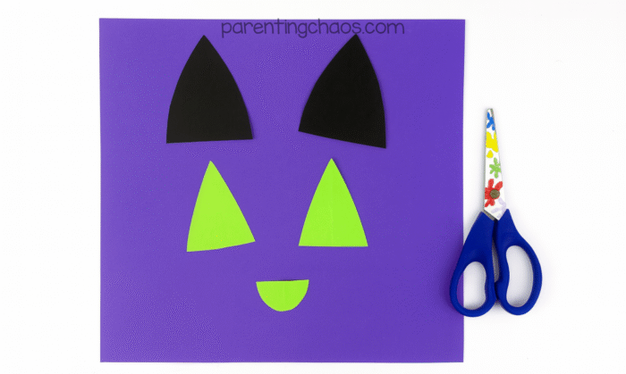 Cut-out shapes for the cat’s ears and nose in black and green construction paper laid out on purple background with scissors to the side.
