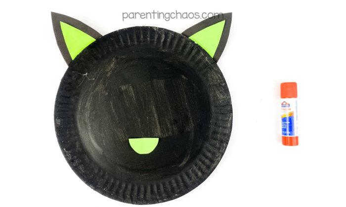 Painted black paper plate with green and black paper ears and a green nose glued on, with a glue stick to the right.