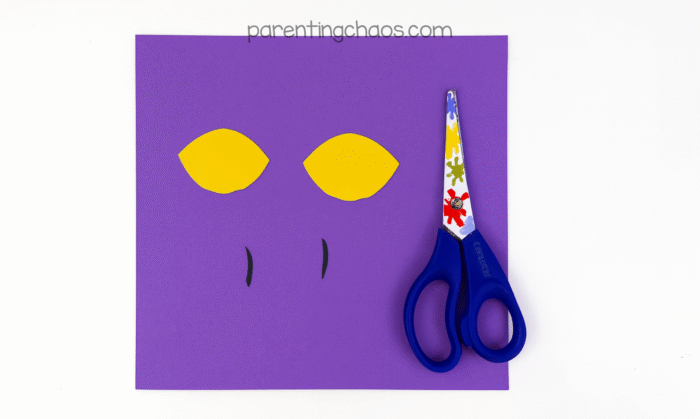 Yellow paper eyes and thin black slits for pupils cut out and placed on a purple background next to scissors.