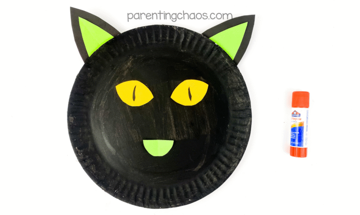 Black paper plate with attached green ears and yellow eyes with black pupils, plus a green paper nose, ready for whiskers.
