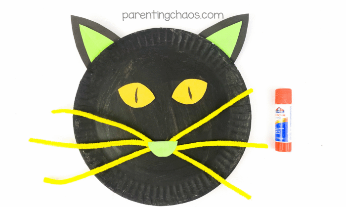 Black cat paper plate craft with yellow pipe cleaner whiskers glued on, yellow eyes, green nose, and ears attached, with glue stick on the right.