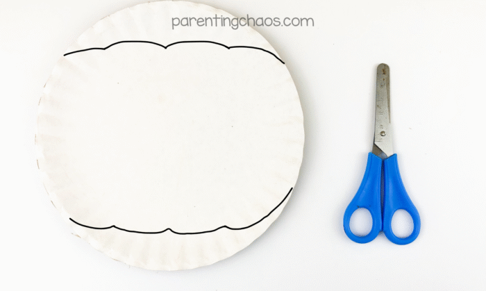 A white paper plate with a curved pumpkin shape drawn in black marker, next to a pair of child-safe scissors ready for cutting.