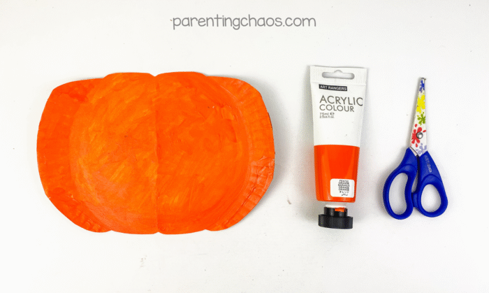 A paper plate painted bright orange, shaped like a pumpkin, with a pair of scissors and orange acrylic paint beside it.