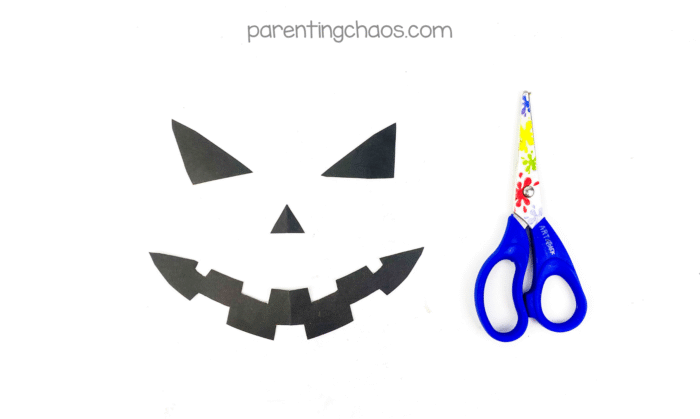 Cut-out black paper shapes for the Jack-o’-Lantern’s face — two triangle eyes, a small triangle nose, and a jagged toothy smile — next to scissors.