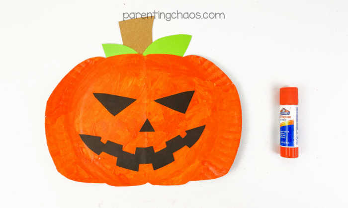 The paper plate pumpkin now has its spooky black paper face glued on, forming a classic grinning Jack-o’-Lantern.