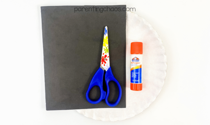 Black construction paper, preschool scissors with colorful handles, a glue stick, and a white paper plate laid out on a white surface for a skull craft.