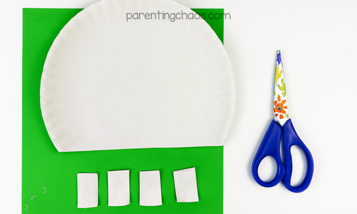 Paper plate with the bottom trimmed flat and five rectangular pieces cut out, placed on a bright green sheet of paper next to preschool scissors.