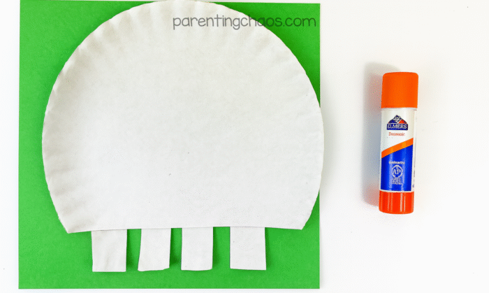 Paper plate with five rectangular teeth glued to the bottom edge, sitting on a green background with a glue stick to the right.