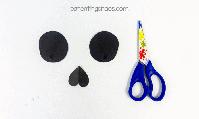 Two large black circles for eyes and a black upside-down heart shape for the nose cut from construction paper, with preschool scissors beside them.