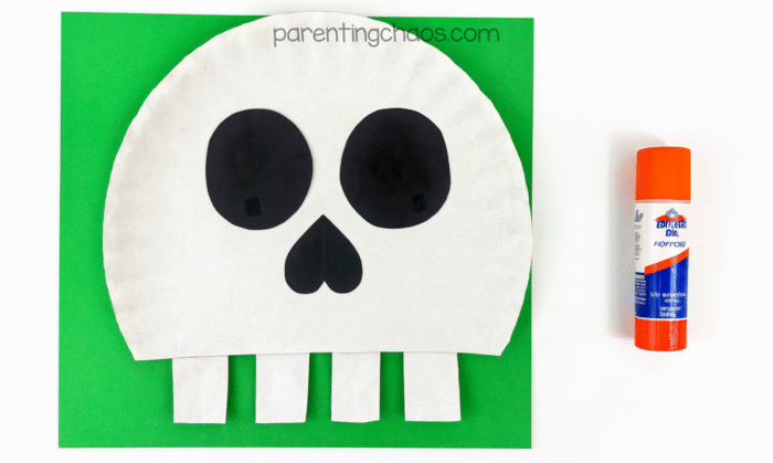 Completed skull craft with black eyes and a heart-shaped nose glued onto a paper plate with teeth at the bottom, set against a green background with a glue stick.