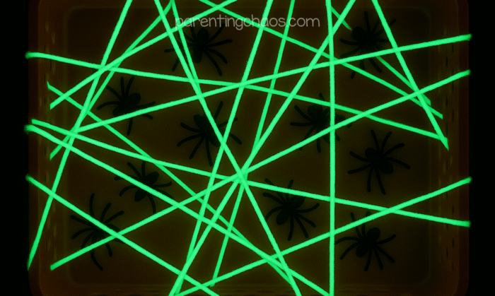 Glow-in-the-dark spider web sensory bin with black plastic spiders tangled under bright green yarn.