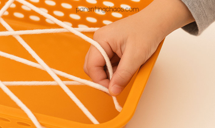 A child’s hand weaving white yarn through an orange sensory tray to create a spider web for a fine motor Halloween activity.