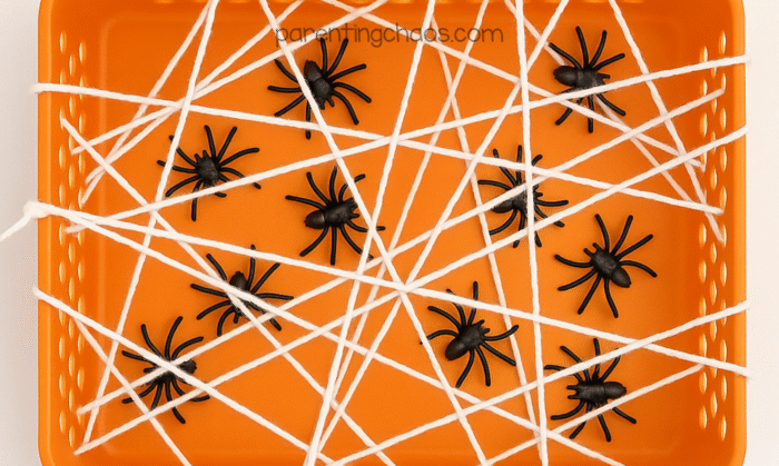 Halloween sensory bin with white yarn woven like a spider web over an orange tray, black spiders scattered underneath.
