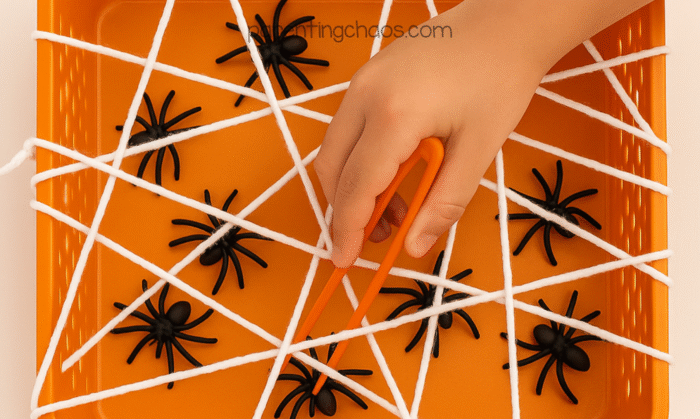 Child using orange tongs to grab black plastic spiders through the yarn web in a Halloween sensory bin activity.