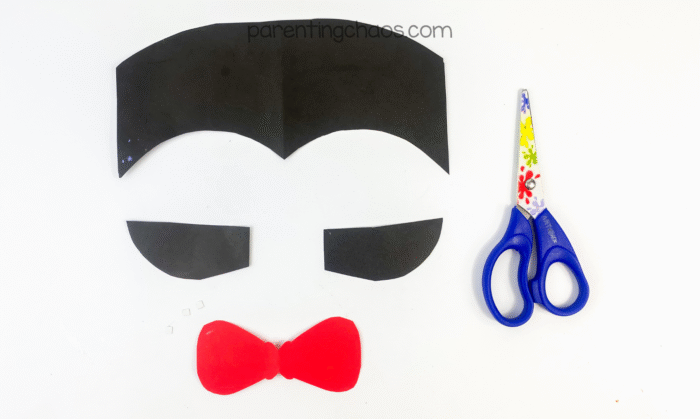 Cut pieces of black construction paper for the vampire’s hair and ears, and a red bow tie, with scissors beside them.