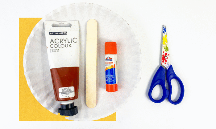 Supplies for witch broom paper plate craft — brown acrylic paint, glue stick, craft stick, scissors, and paper plate.