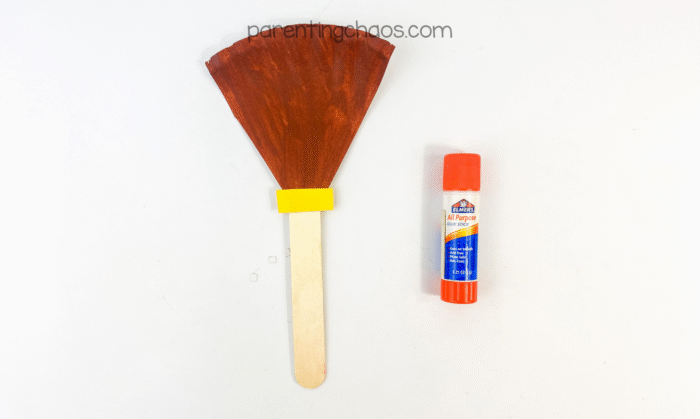 Brown paper plate broom top glued to craft stick with yellow band.