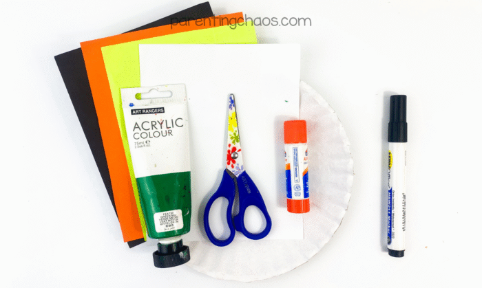 Overhead view of witch paper plate craft supplies including green acrylic paint, glue stick, scissors, marker, paper plate, and construction paper.