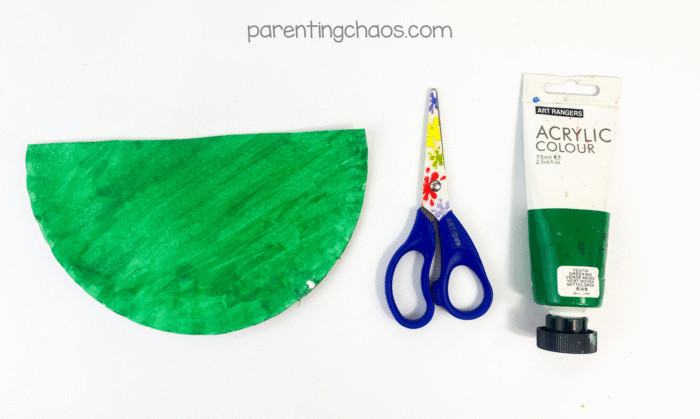 Half-painted paper plate painted green next to scissors and a tube of green acrylic paint.