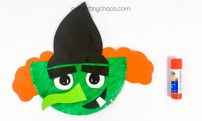 Assembled witch craft with hat, orange hair, big eyes, and green face beside a glue stick.