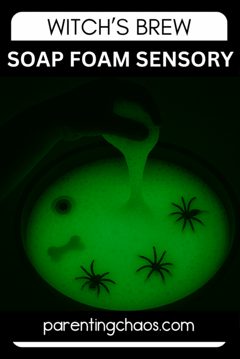 Hand lifting glowing green soap foam from a witches brew sensory bin with toy spiders.