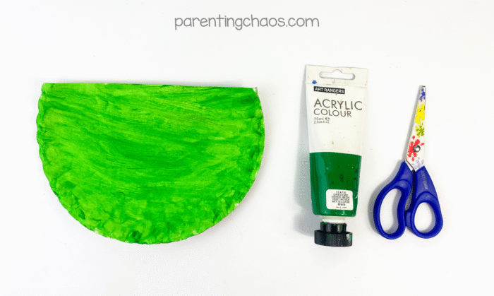 three-fourths paper plate painted bright green with paint tube and scissors beside it.