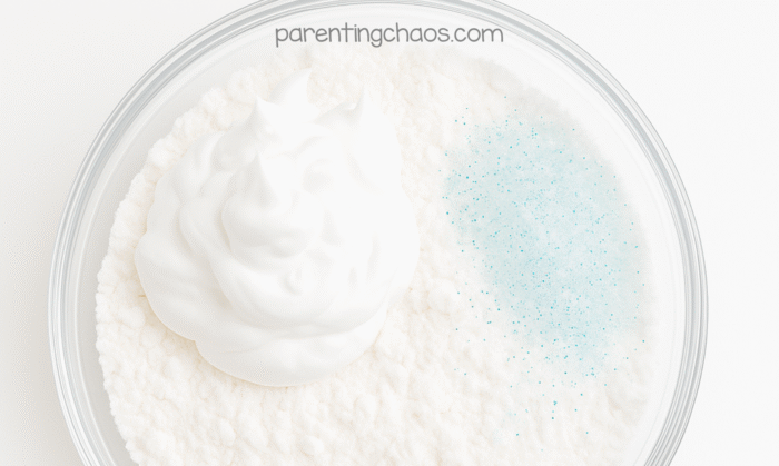 Close-up of a clear bowl filled with baking soda, a mound of shaving cream, and a sprinkle of iridescent glitter before mixing to make fake snow.