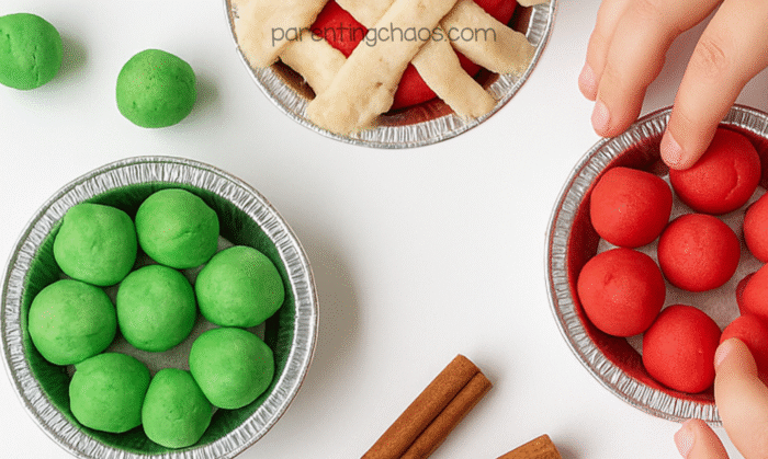 Mini pie tins filled with red and green apple pie playdough balls, with a child’s hand pressing one into place.