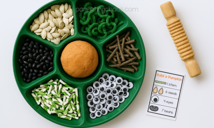 Overhead view of a green tray filled with playdough tray materials for a pumpkin activity, including orange playdough, pumpkin seeds, sticks, black beans, pipe cleaners, paper straws, and googly eyes, with a wooden rolling pin and instruction card beside it.