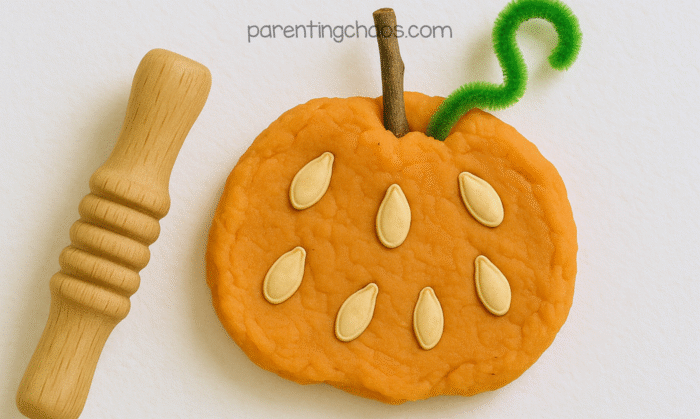 Close-up of a finished orange playdough pumpkin decorated with pumpkin seeds, a stick stem, and a green pipe cleaner vine next to a wooden rolling pin.