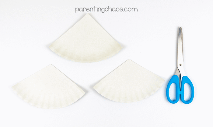 Three white paper plates cut into large triangle sections beside scissors, ready for the paper plate Christmas tree craft for kids.