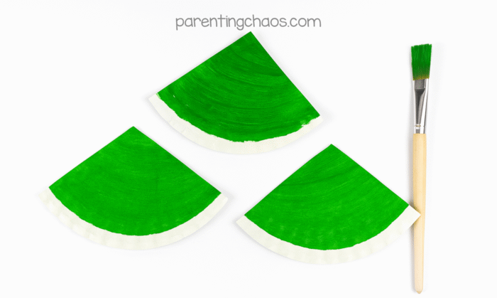 Triangular paper plate pieces painted bright green with a white edge, drying next to a paintbrush for the paper plate Christmas tree craft for kids.