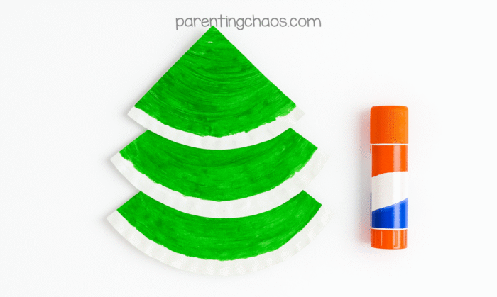 Green painted paper plate sections stacked and glued into a tree shape with a glue stick beside them, part of the paper plate Christmas tree craft for kids.