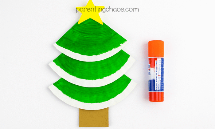 Completed tree shape made from painted paper plate pieces, a brown trunk, and a yellow star on top, next to a glue stick—part of the paper plate Christmas tree craft for kids.